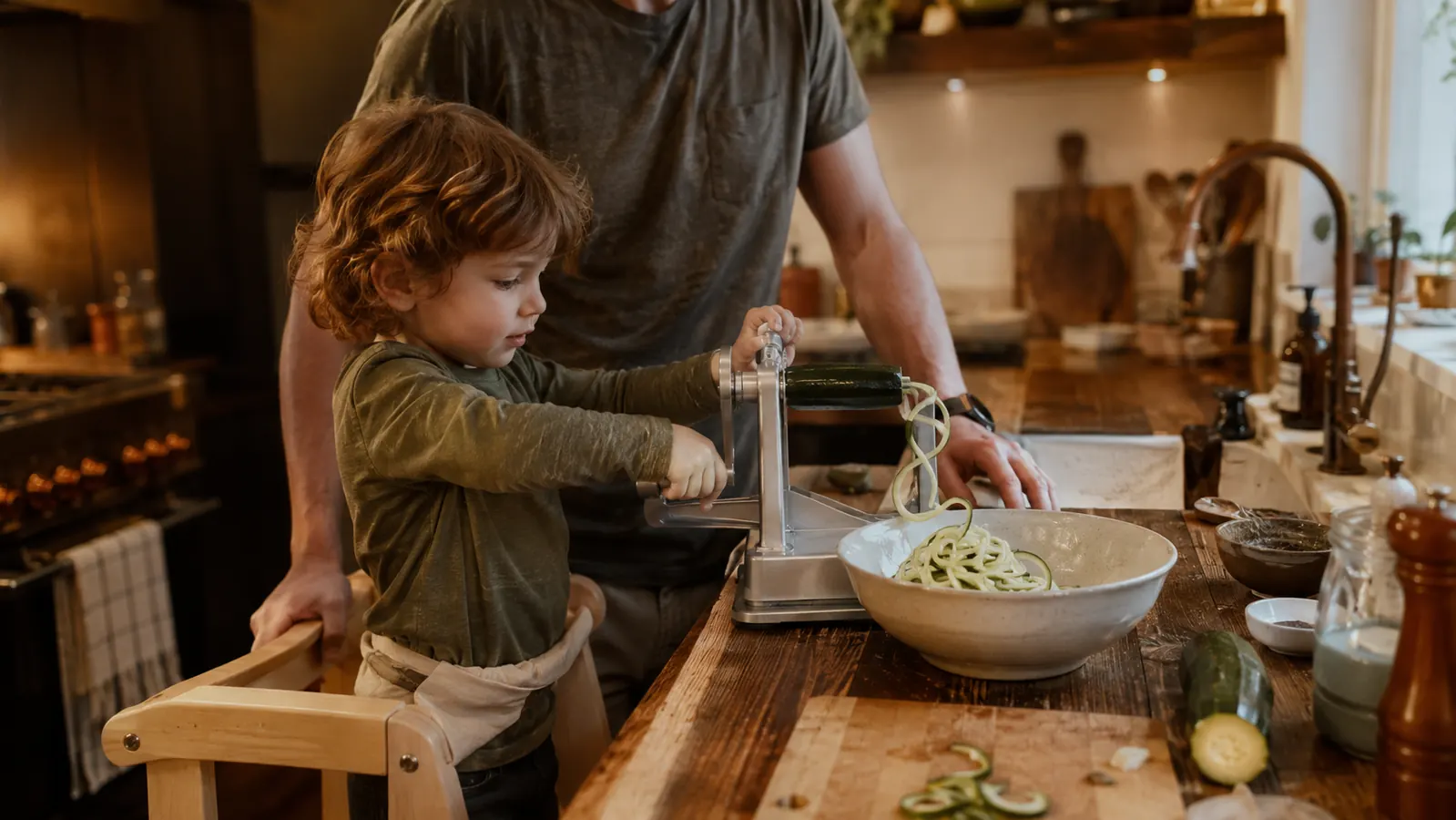 A small child standing on a step stool at a kitchen counter reaching for a spiralizer handle