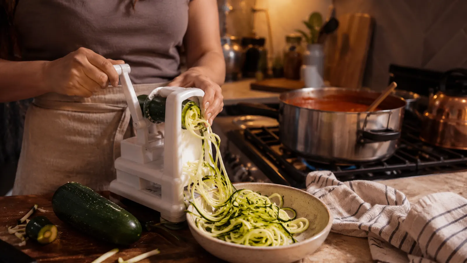 A kitchen counter at dinnertime with a bowl of spiralized zucchini noodles next to a spiralizer and a pot of marinara